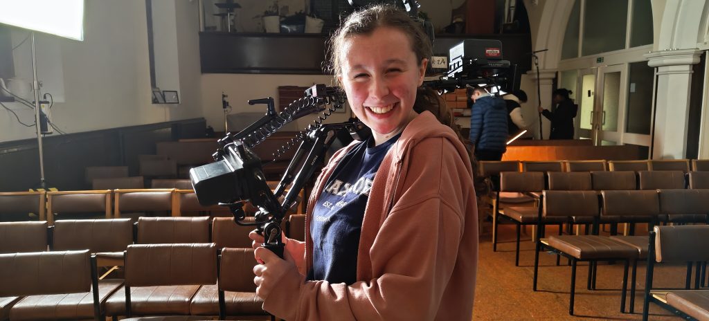 A young Film Academy camera operator smiling as she holds a rigged camera in a theatre auditorium.
